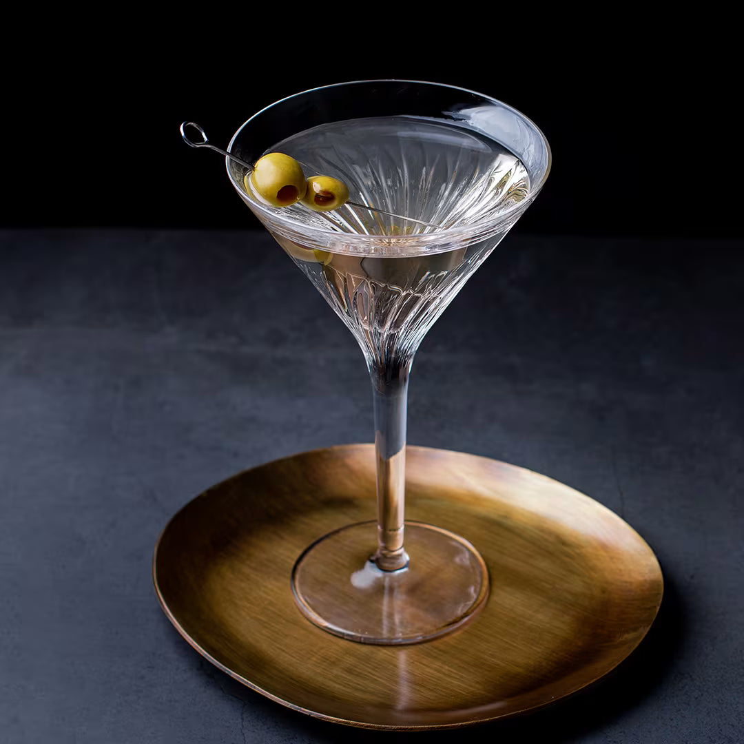 A clear cut-glass martini coupe filled with a transparent cocktail and garnished with two olives on a skewer, set on a round brushed gold plate against a dark studio background.