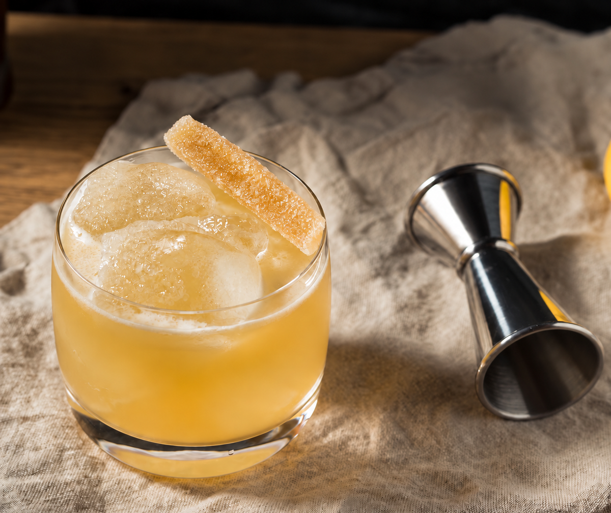 A clear rocks glass filled with an amber-yellow cocktail and large ice cubes, garnished with a sugar-skinned orange peel, set on a beige linen cloth beside a stainless steel jigger.