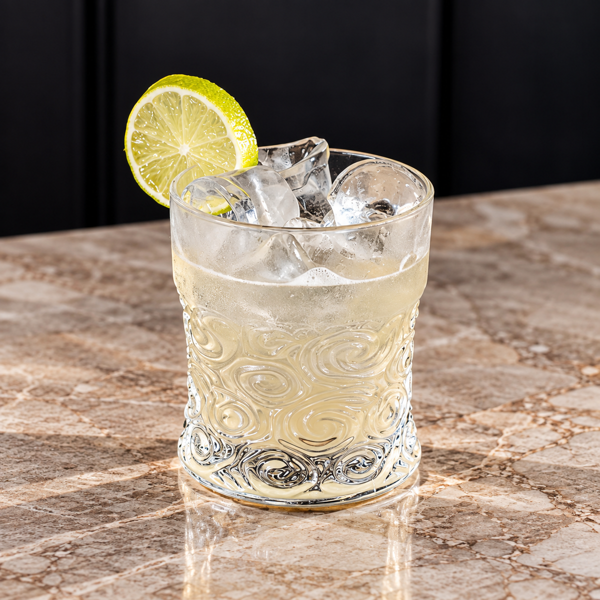 A clear textured rocks glass filled with a pale yellow drink and ice, garnished with a lime slice, set on a marble countertop against a dark background.