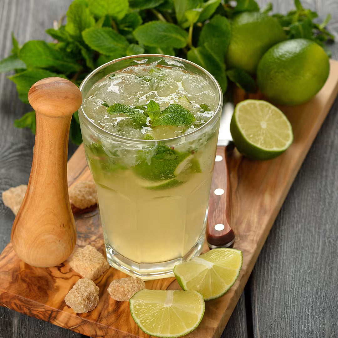 A clear glass filled with a pale yellow iced beverage, mint leaves, and lime slices, set on a wooden cutting board with limes, a wooden muddler, sugar cubes, and a knife.